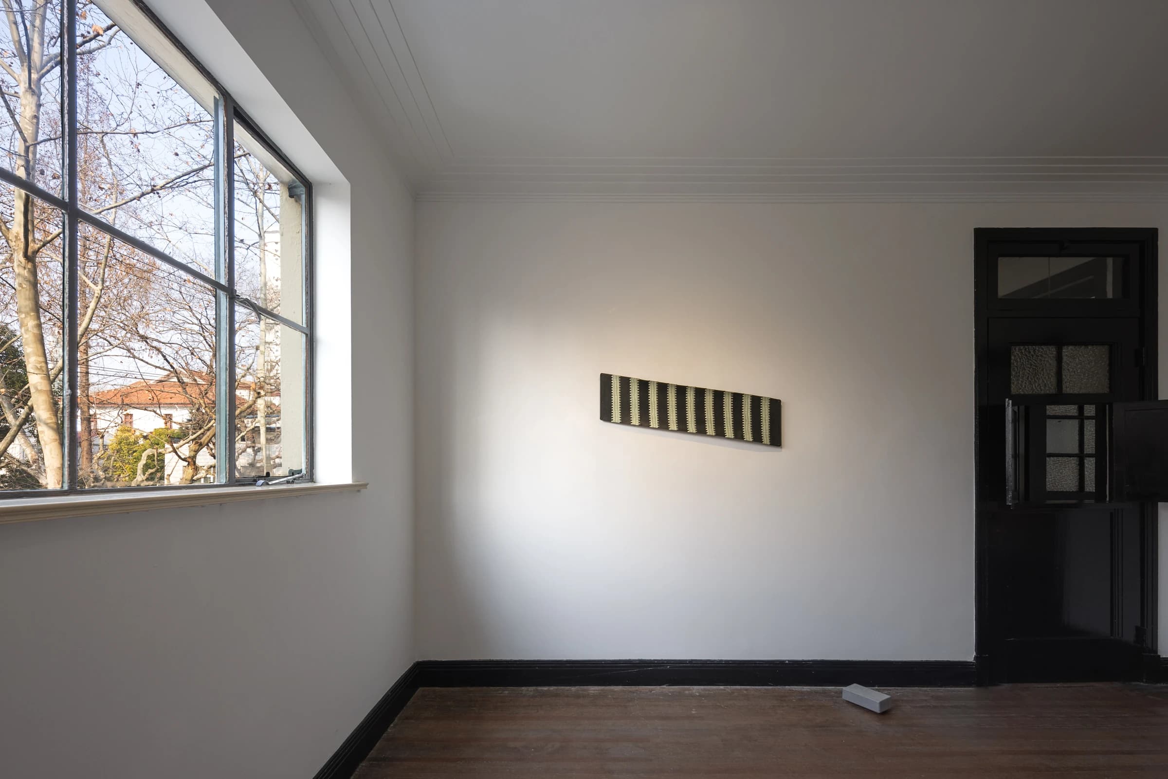 白色展間中,橫幅畫作掛在窗邊,自然光映照木地板與作品。